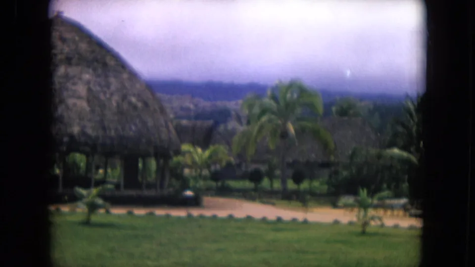 1968 Honolulu resort view with a thatched cabana at left, palms, lawn, paths, and distant hills in soft 8mm focus.