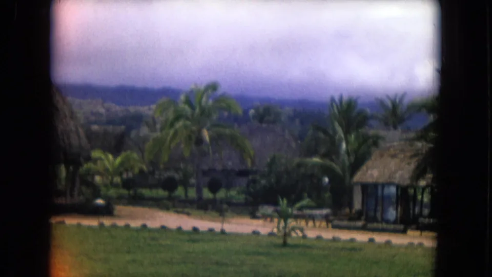 1968 Honolulu home-movie view across resort lawns toward palms, low thatched structures, paths, and hazy hills.