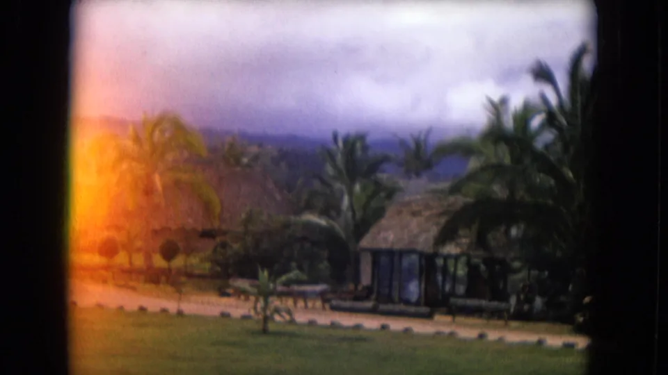 1968 Honolulu resort frame showing palms, thatched roofs, lawn, and a warm film flare along the left side.