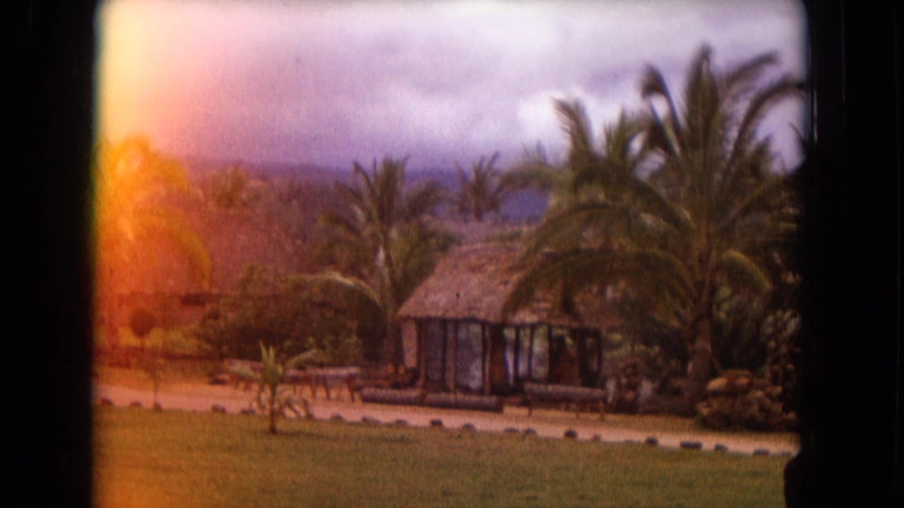 1968 Honolulu home-movie frame showing a thatched hut, palms, grass, and water beyond the landscaping.