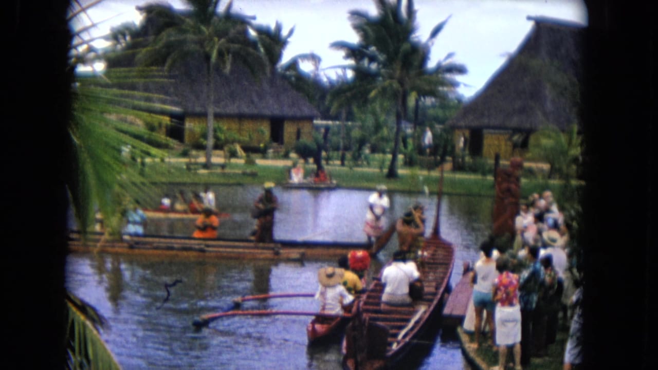 Tourists watch outrigger canoes on a lagoon beside thatched buildings and palm trees.