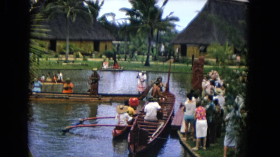 1968 Honolulu frame with tourists on the dock, canoe riders, performers in costume, carved posts, palms, and thatched-roof buildings.