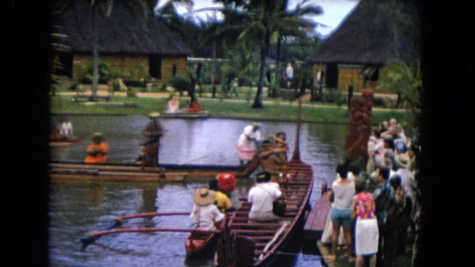 Home-movie frame from 1968 Honolulu showing a performer, outrigger canoes, tourists, a dock, lagoon water, palms, and thatched structures.