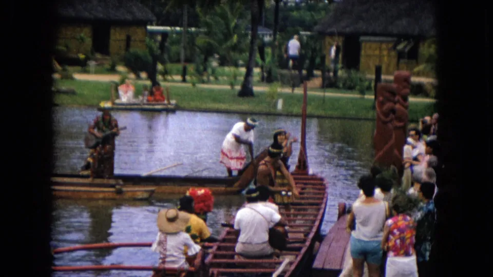 1968 Honolulu frame showing a performer stepping near an outrigger canoe, tourists on a dock, lagoon water, palms, and thatched buildings.