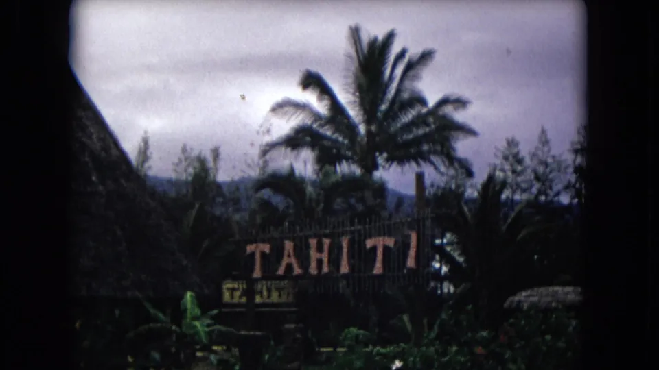 1968 Honolulu home-movie frame showing a Tahiti sign partly framed by a thatched roof, palm trees, foliage, and distant hills.