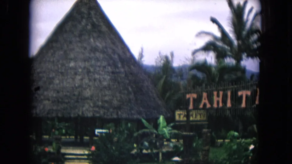 1968 Honolulu frame with a large conical thatched hut, partial Tahiti sign, palm trees, paths, and tropical garden foliage.