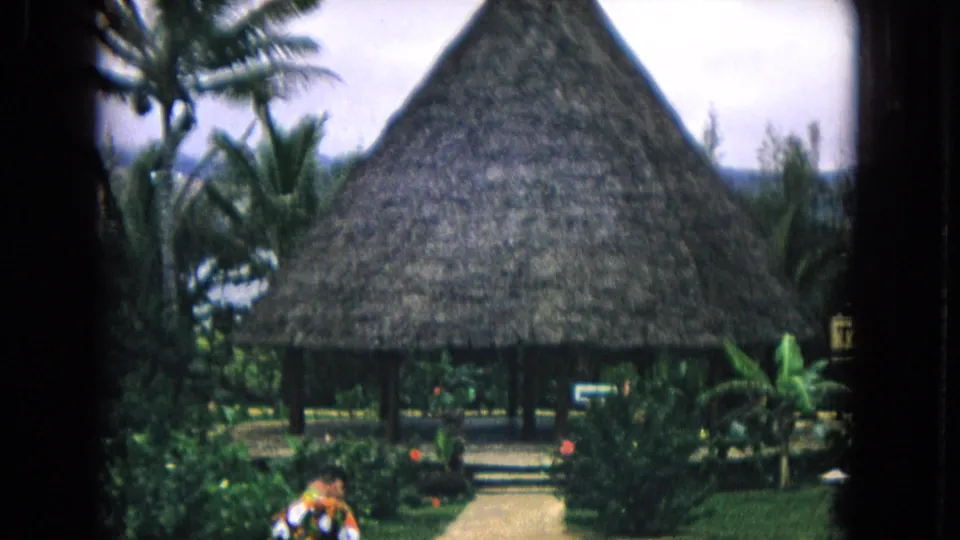 1968 Honolulu frame with a thatched building in tropical landscaping and a person partly visible near the foreground path.