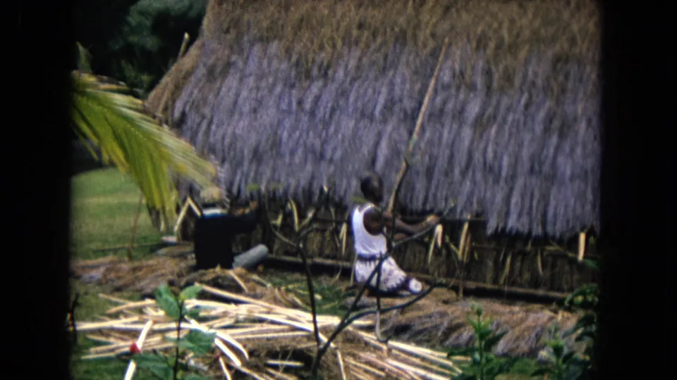 1968 Honolulu 8mm frame showing a seated person near a thatched structure as another figure works with a long pole outdoors.