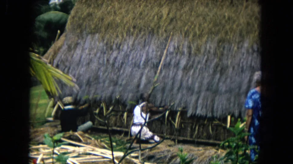 Honolulu 1968 home-movie frame of a seated figure near a thatched wall, with tools or poles and a second person partly visible.