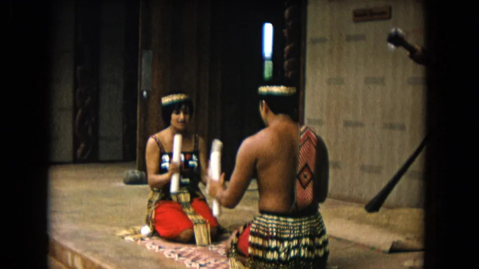 1968 Honolulu 8mm frame of two traditionally dressed seated performers facing each other on a mat in a staged interior or sheltered setting.