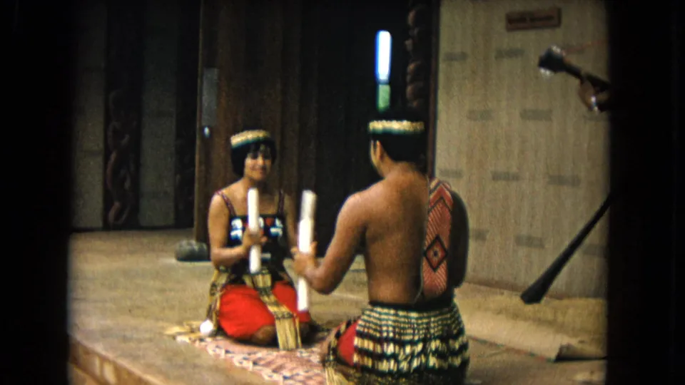 Honolulu 1968 frame showing two seated performers in traditional dress holding objects, with a dark pole or prop visible at frame right.