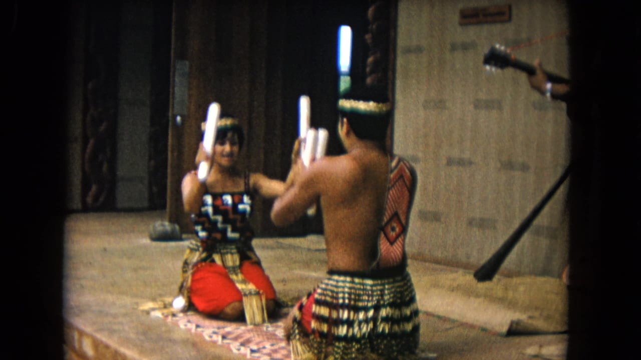 Two seated adult performers face each other while holding white rhythm sticks indoors.