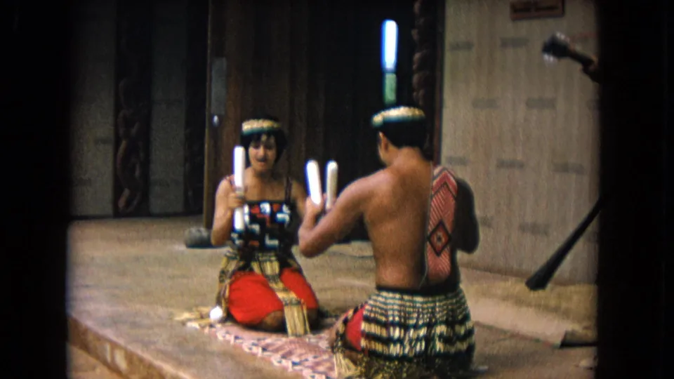 A man and woman continue the seated stick rhythm routine in 1968 Honolulu footage, framed by warm wood walls and carved posts.