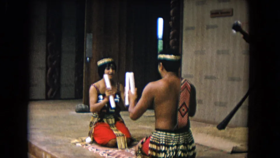 In the final frame, the performers hold white sticks upright while seated on a mat in the 1968 Honolulu indoor performance scene.