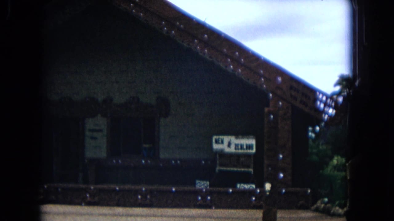 1968 Honolulu home-movie frame showing a carved meeting-house exterior with a dark entrance, posted notices, and a New Zealand sign.