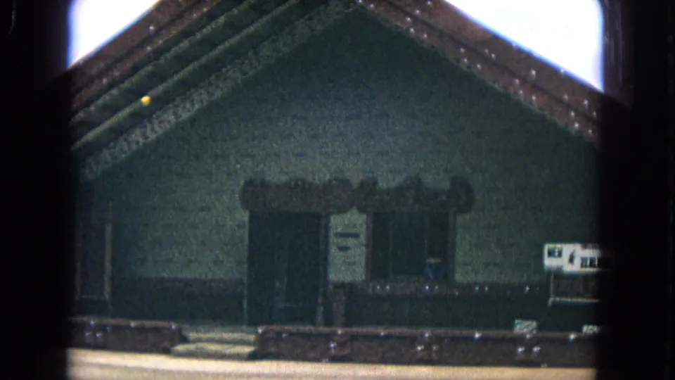 Closer 1968 Honolulu home-movie view of the carved meeting-house front, including the doorway, posted notices, low barrier, and partial New Zealand sign.