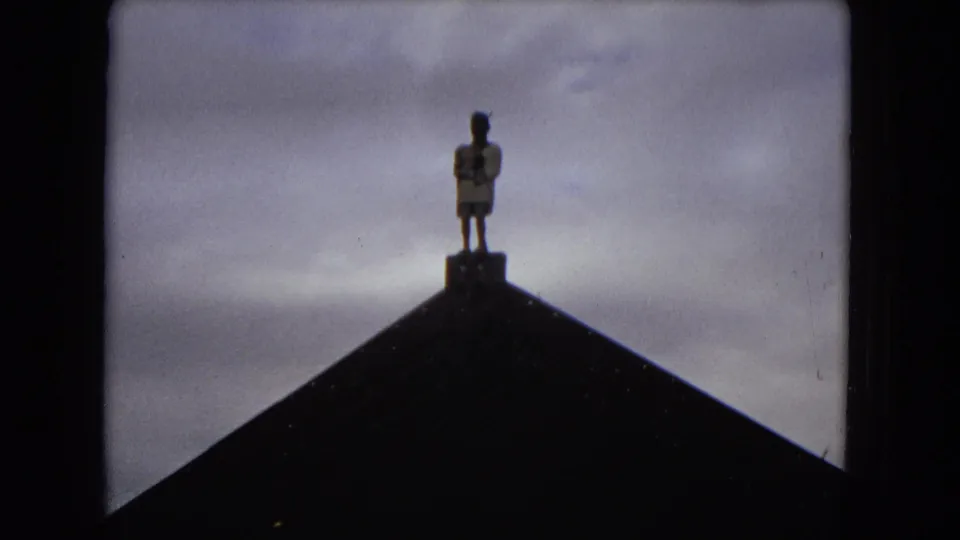 1968 Honolulu 8mm frame with a silhouetted figure on the peak of a dark roof, framed against a cloudy pale sky.