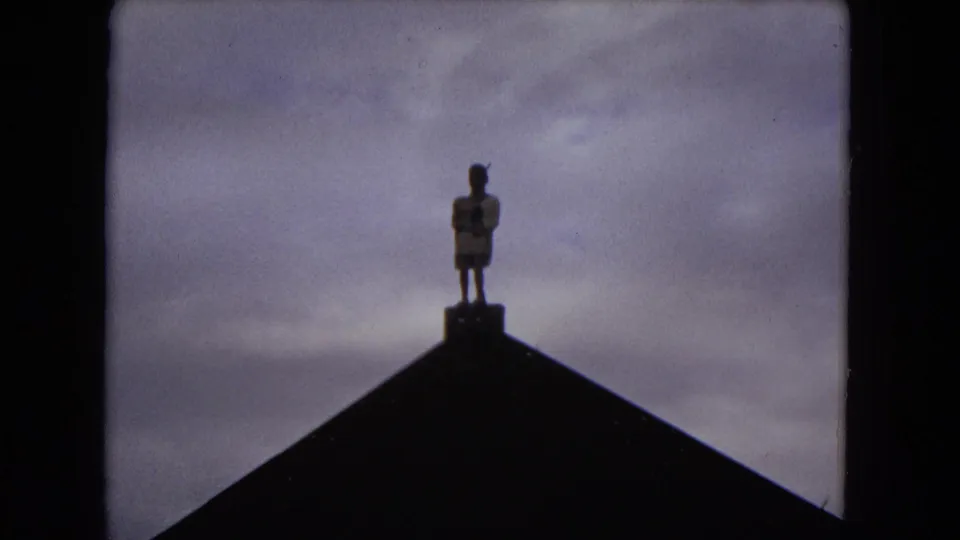 1968 Honolulu 8mm frame showing a small figure or statue silhouetted on a peaked roofline beneath a muted sky.