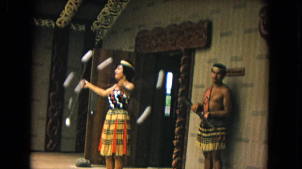 1968 Honolulu 8mm frame of a woman twirling poi balls in a grass skirt as a male performer stands inside a carved performance interior.