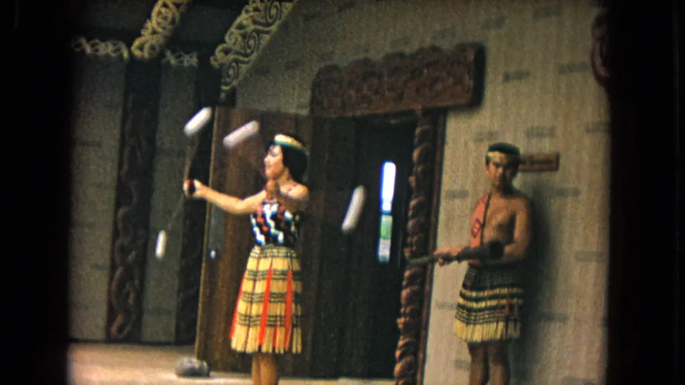1968 Honolulu 8mm frame showing a woman in patterned top and grass skirt spinning poi balls beside carved interior posts.