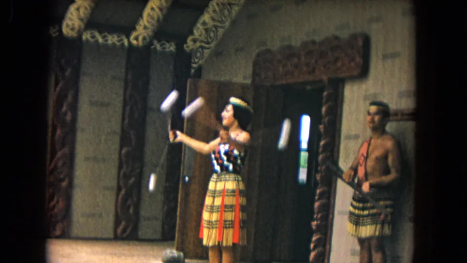 1968 Honolulu 8mm frame of a poi performance with a woman in costume foregrounded and a male performer near a carved hut doorway.