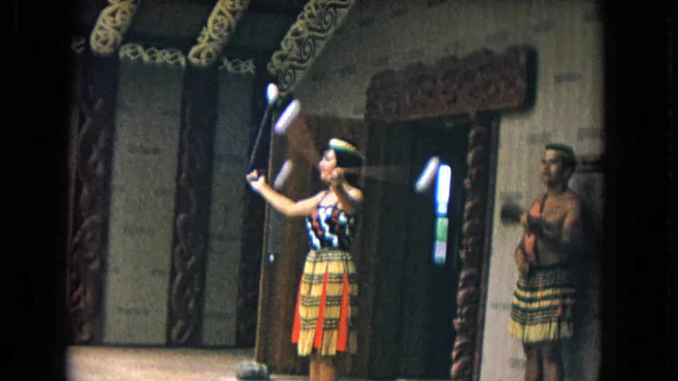 1968 Honolulu home-movie frame showing a female performer with clubs beside a male performer in a carved wooden performance setting.