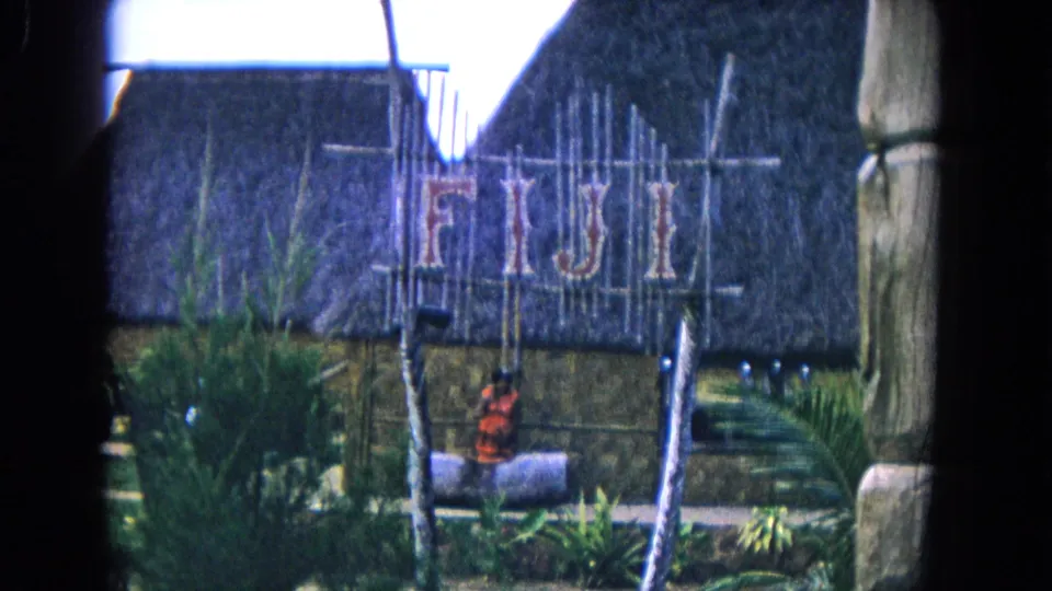 1968 Honolulu home-movie view of a Fiji sign mounted before a thatched hut display with greenery in the foreground.