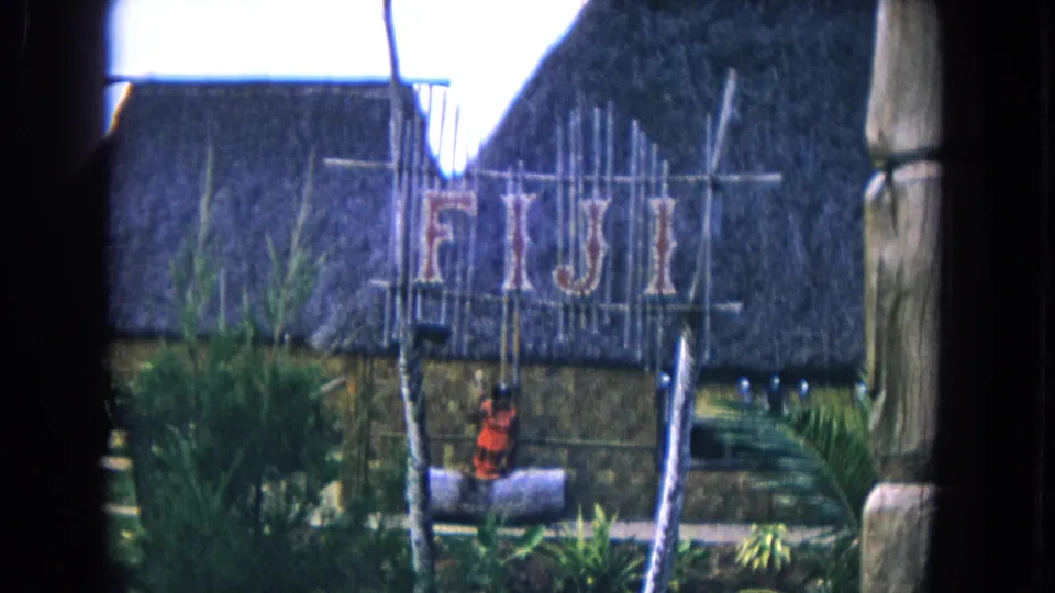 1968 Honolulu 8mm frame of a Fiji-themed thatched display, with the sign centered and tropical plants below.