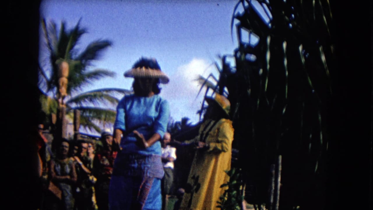 Woman in blue performing hula beside a dancer in yellow as spectators sit behind them outdoors.
