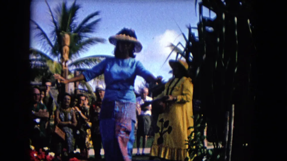 1968 Honolulu outdoor hula scene showing the blue-dressed dancer gesturing beside a yellow-dressed dancer and watching crowd.