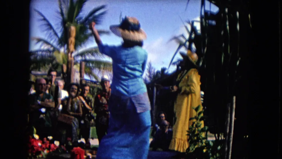 1968 Honolulu hula performance from a rear angle, with the blue-dressed dancer raising an arm toward another dancer and spectators.
