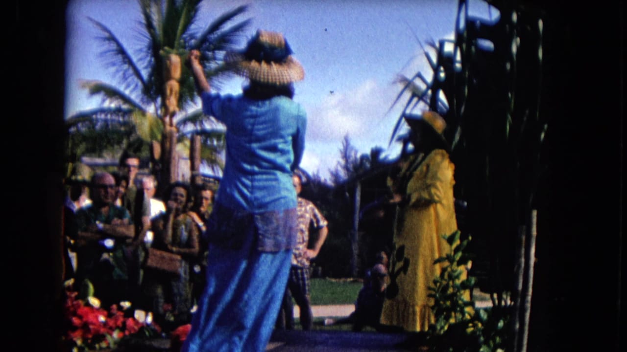 Woman in a blue dress dances hula before tourists in an outdoor palm-lined setting.