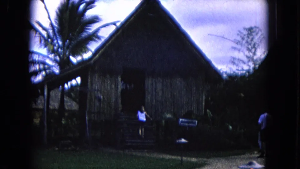 1968 Honolulu home-movie view of a thatched performance structure with palms, visitors, and a small sign.