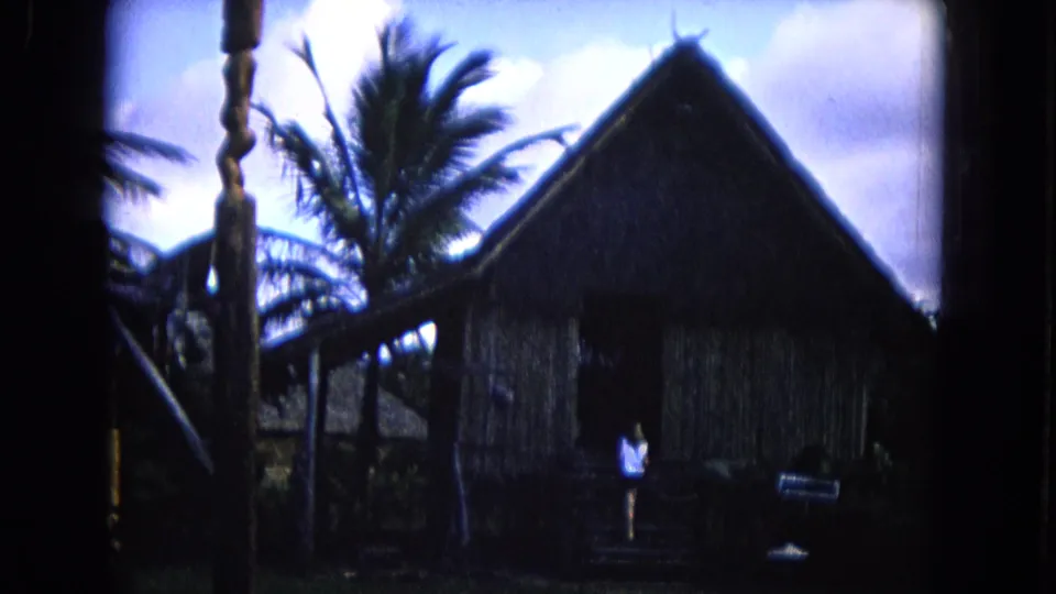 1968 Honolulu frame with a palm-lined thatched structure, stage post, small sign, and a visitor in the foreground area.