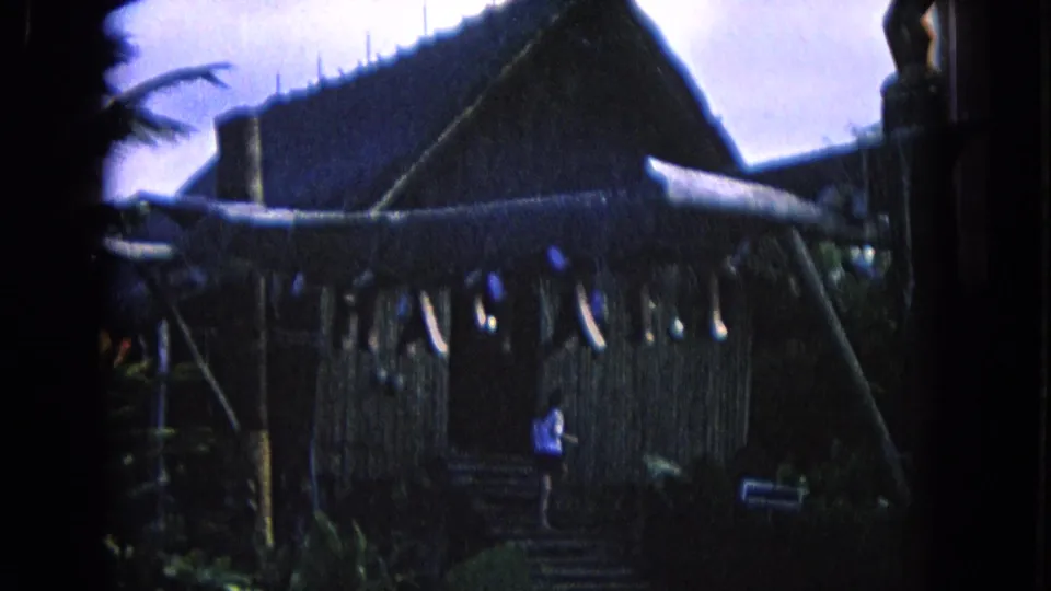 1968 Honolulu home-movie frame showing a thatched structure, railing, foliage, and a person in a light shirt near the entry area.