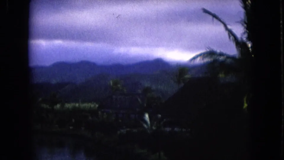 Storm clouds gather over distant Honolulu hills, with palm silhouettes and tropical vegetation below.
