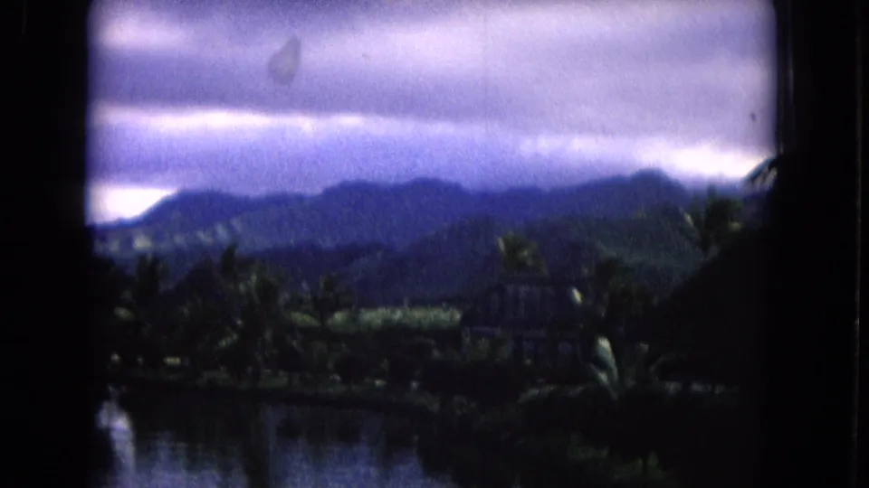 A 1968 Honolulu landscape frame shows rain-heavy clouds over hills, palms, and water in soft 8mm color.