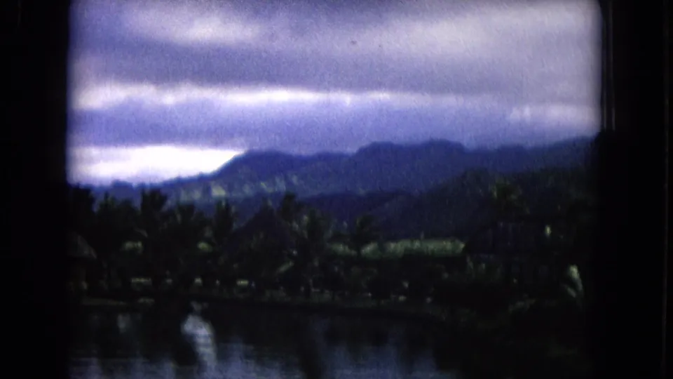 Moody Honolulu home-movie frame of dark clouds, distant hills, palms, and water as rain approaches.