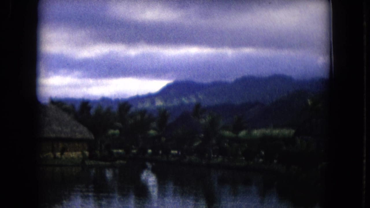 Calm water reflecting a thatched-roof hut, palms, mountains, and cloudy sky.