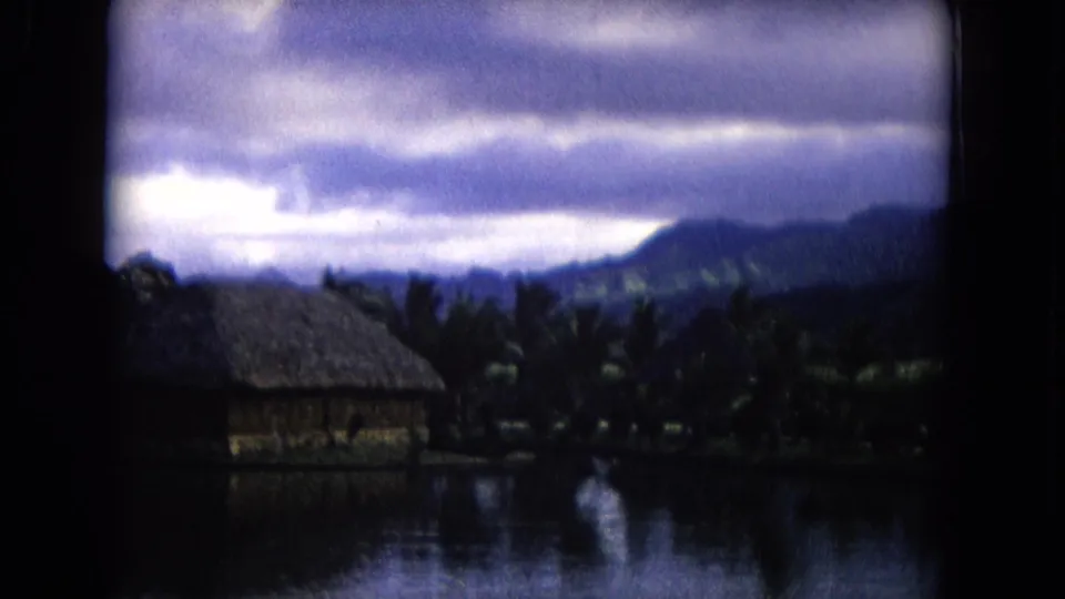 1968 Honolulu home-movie view of a tropical waterside hut, palms, reflective water, and mountain ridges beneath bright clouds.