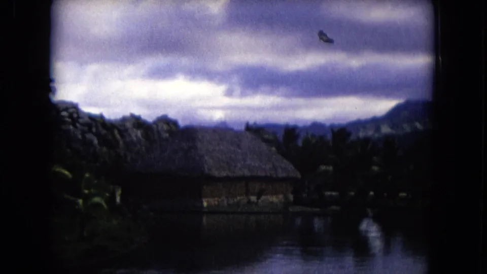 1968 Honolulu frame with a larger thatched hut across calm water, dark palms, distant ridges, and soft overcast sky.