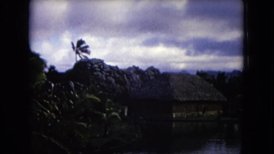 1968 Honolulu 8mm frame showing a thatched waterside hut framed by palms and dark foliage, with mountains and clouds behind.