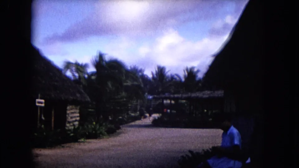 1968 Honolulu home-movie frame showing thatched structures, a path, tropical plants, and a distant person in blue at right.