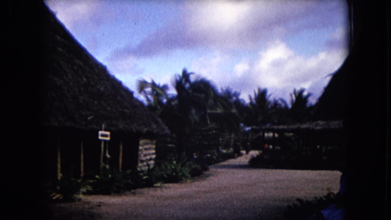 1968 Honolulu 8mm frame showing thatched-roof huts along a quiet path under a cloudy tropical sky.