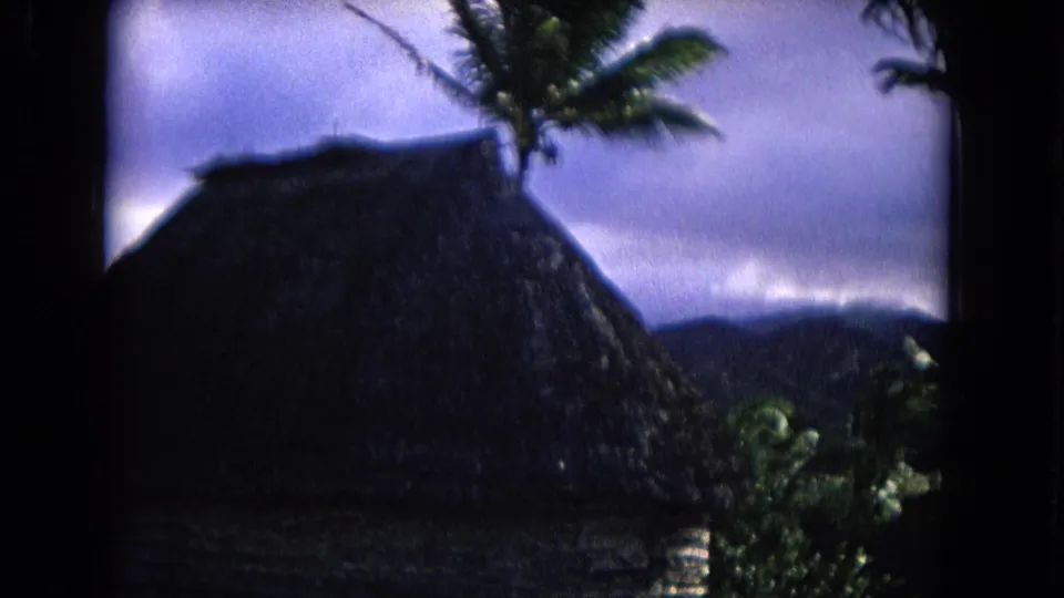 1968 Honolulu 8mm frame showing a hut roof, palm fronds, vegetation, and distant hills under clouds.