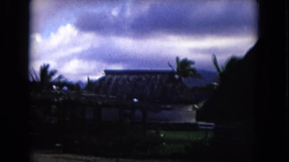 1968 Honolulu-area home-movie frame with palms, a thatched building, cloudy sky, and strong shadowed frame edges.