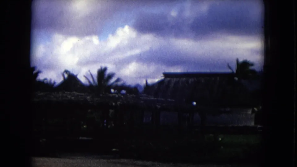 1968 Honolulu 8mm frame showing cloudy sky, palm silhouettes, thatched rooftops, and darkened tropical grounds.