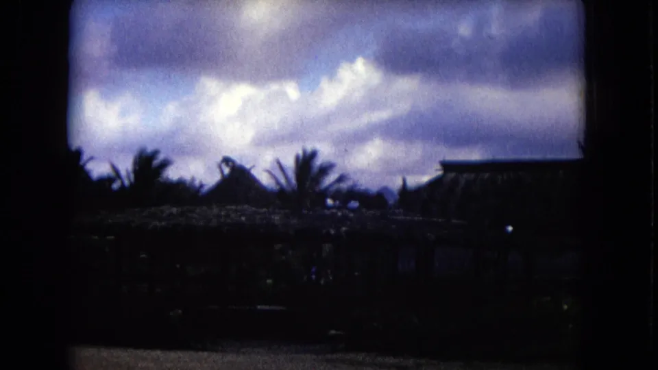 1968 Honolulu home-movie frame with shadowed thatched roofs and palm trees under a bright cloudy sky.