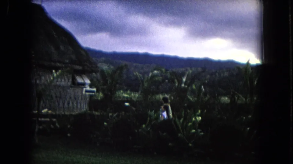 1968 Honolulu 8mm frame showing a tropical hut setting, distant mountains, heavy clouds, palm foliage, and a person in light clothing.