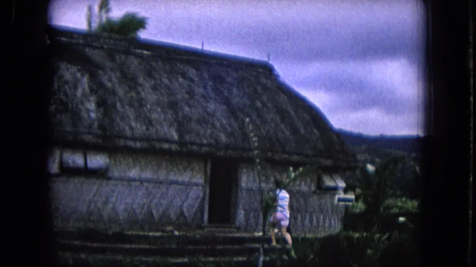 1968 Honolulu frame with a large thatched-roof structure, tropical plants, distant hills, and a person walking beside the hut.
